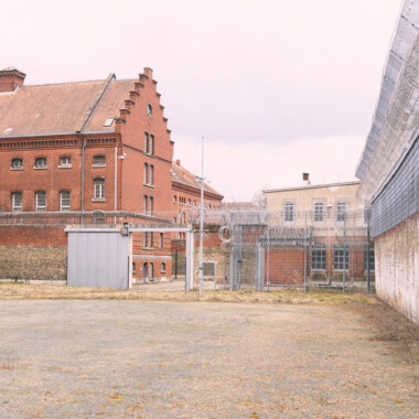 Ein Bild der JVA Rennelberg. Ein rotes Backsteingebäude mit vergitterten Fenstern, einen Hof mit Zäunen und Stacheldraht unter grauem Himmel.